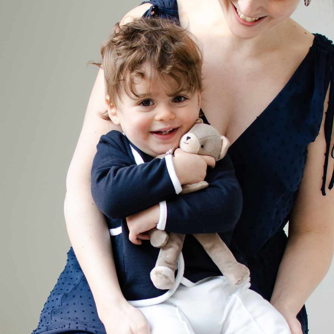 Smiling baby boy with his mother. He is holding on to the Elliott Bear Buddy Pacifier Holder, a floppy stuffed animal bear that clips on to a pacifier.