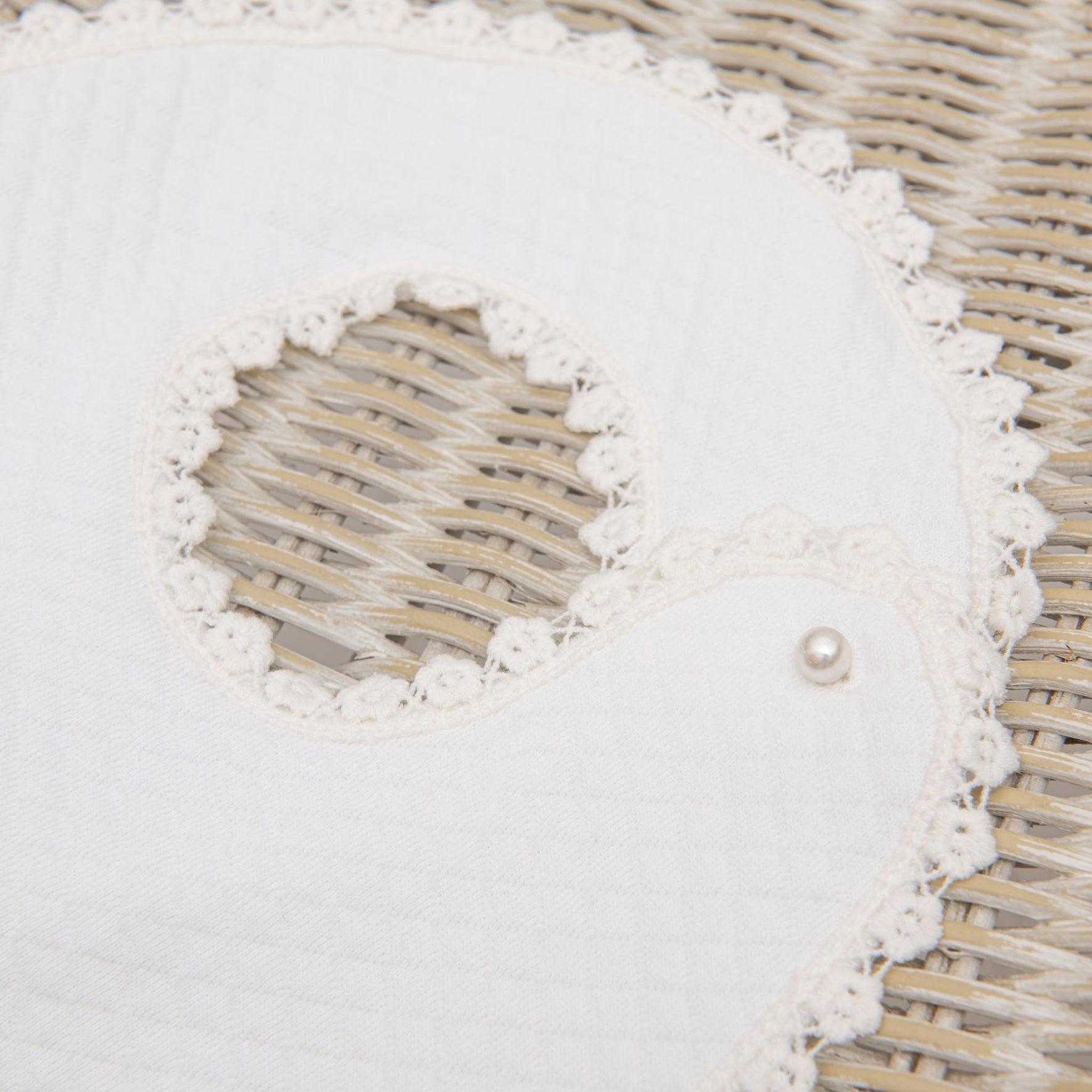 An Eliza Bib, a white, quilted fabric baby bib with crocheted floral lace trim, shaped like a bird, lays on a woven wicker surface. It has a small pearl-like button near the neckline.