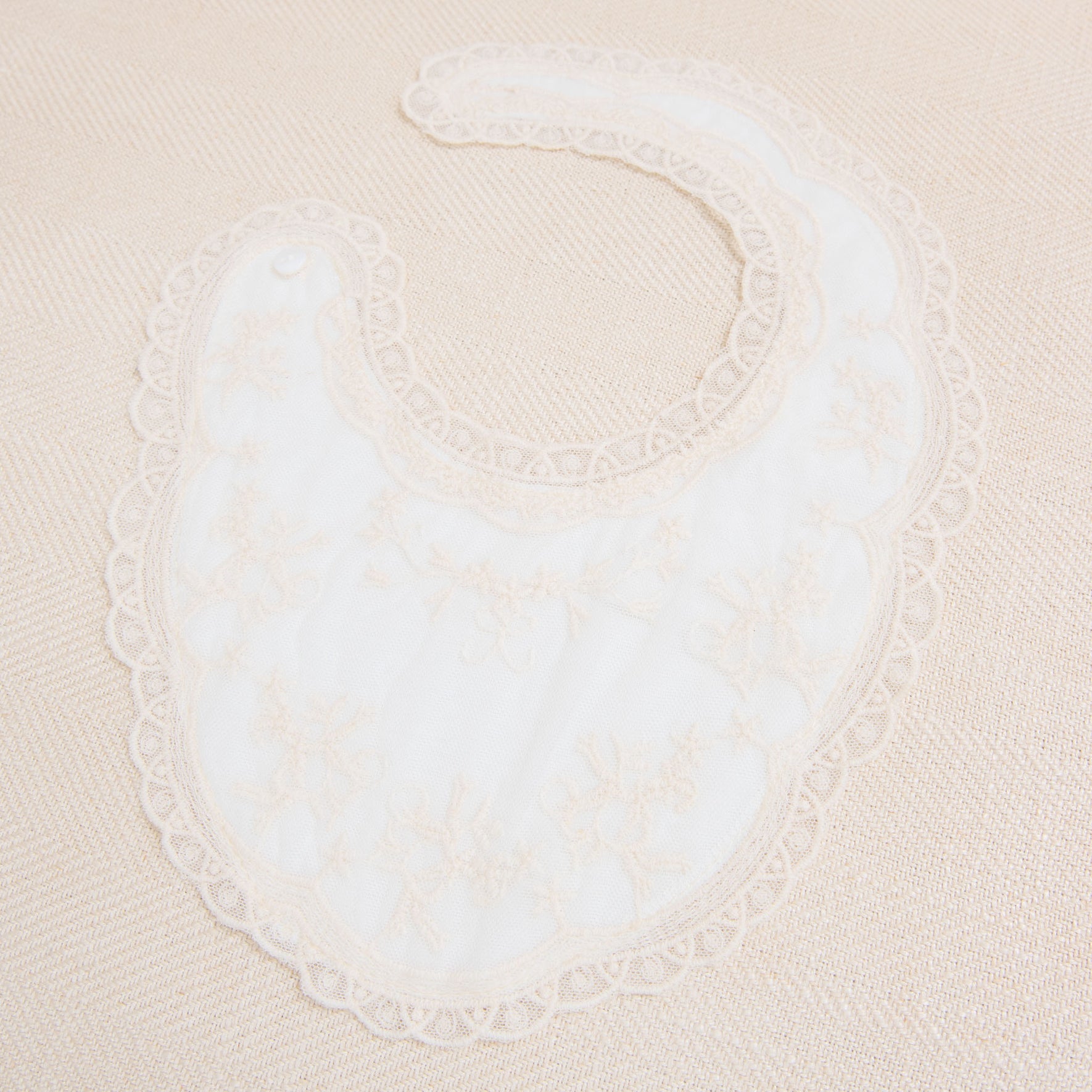 A Jessica Cotton Christening Bib, crafted from soft cotton, is laid out on a beige surface. The full-coverage bib features delicate floral embroidery and a scalloped champagne lace trim. Designed to fasten at the neck with a small snap button, this bib combines elegance with practicality.