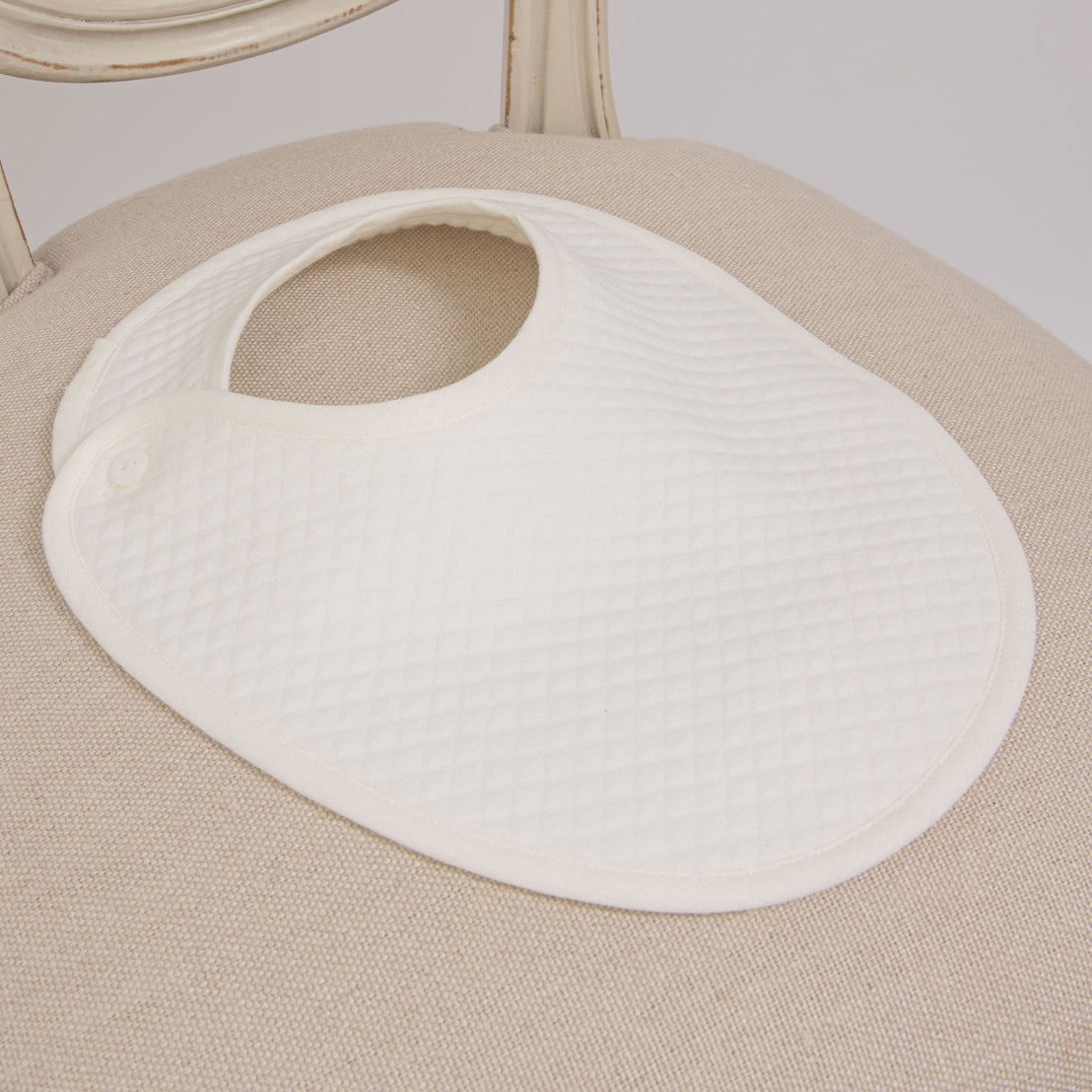 Flat lay photo of the Owen Bib on a chair. The Owen Bib is made out of ivory quilted cotton with an ivory linen trim and button closure.