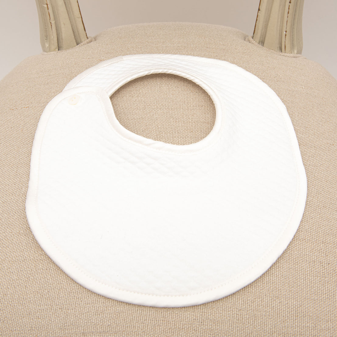 Flat lay photo of the James Bib made from 100% white quilted cotton with button closure and silk piping on the edge.