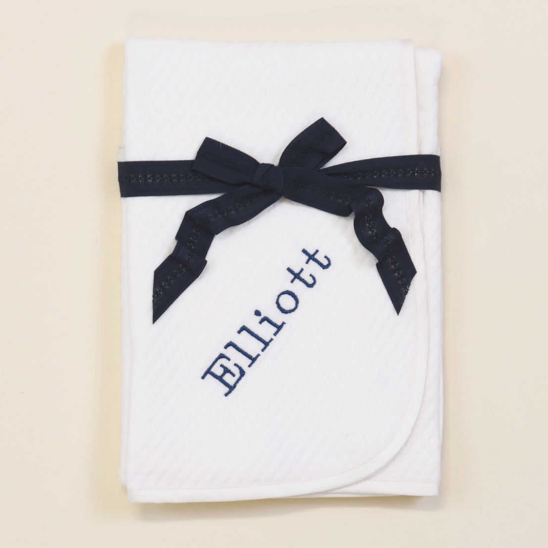 Flat lay photo of the Elliott Personalized Blanket folded up and tied with a navy ribbon. The blanket is made out of a soft white 100% quilted cotton,  one corner has the name "Elliott" embroidered in navy.