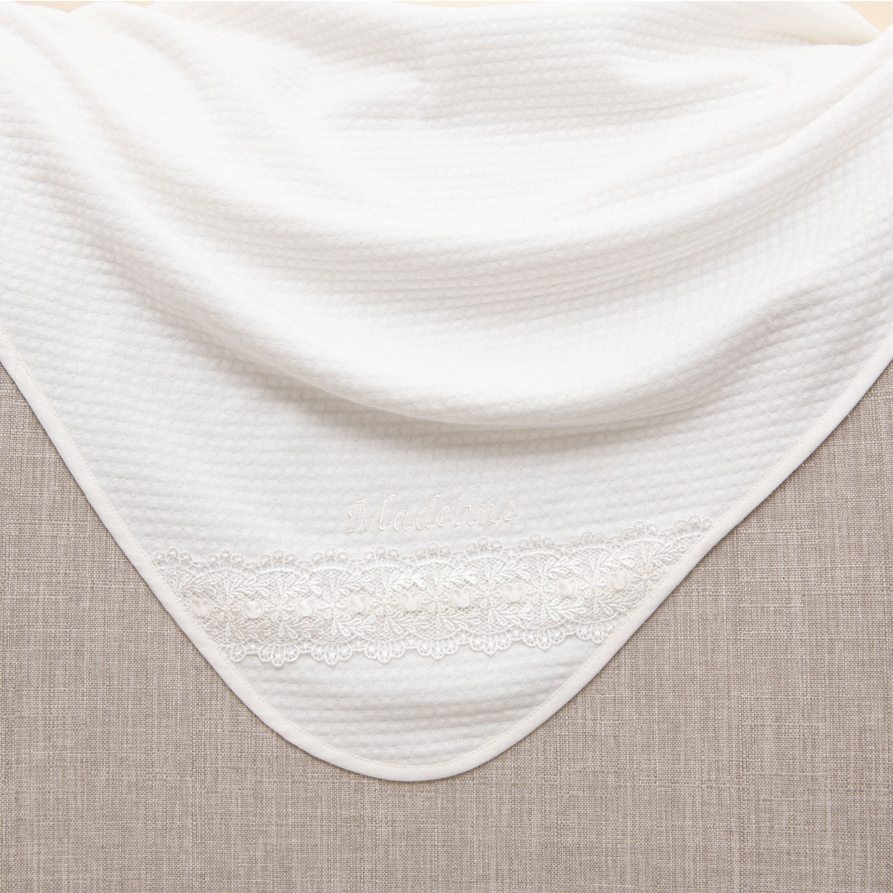 The Madeline Newborn Gift Set - Save 10% includes a white knit blanket showcasing a lace pattern on one end, elegantly draped over a beige surface reminiscent of the gentle hug of a quilted cotton romper. The textured fabric and contrasting colors beautifully highlight the distinct patterns in the material.
