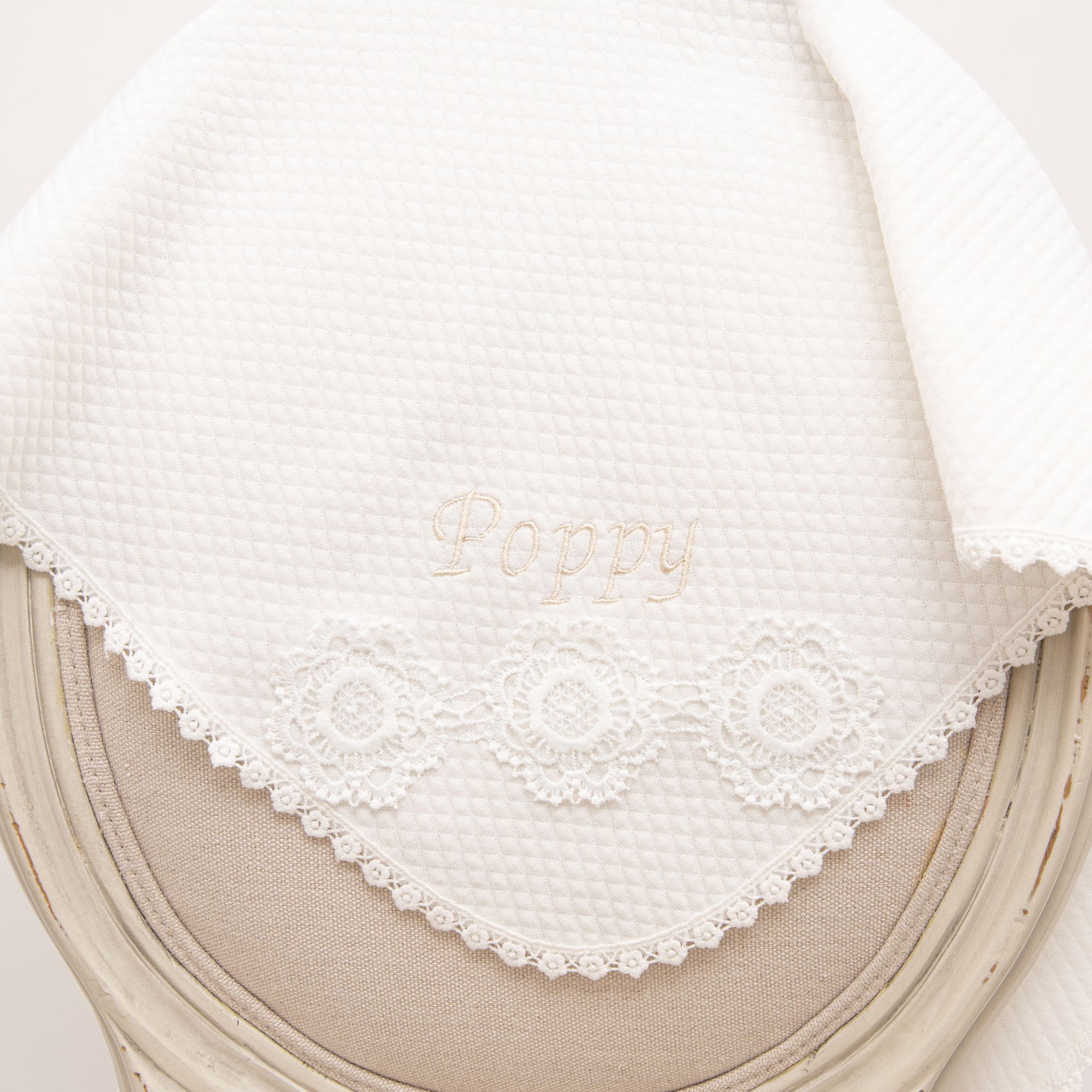 Quilted Poppy Christening Blanket elegantly displayed on a chair, showcasing delicate floral lace trim and 'Poppy' embroidery.