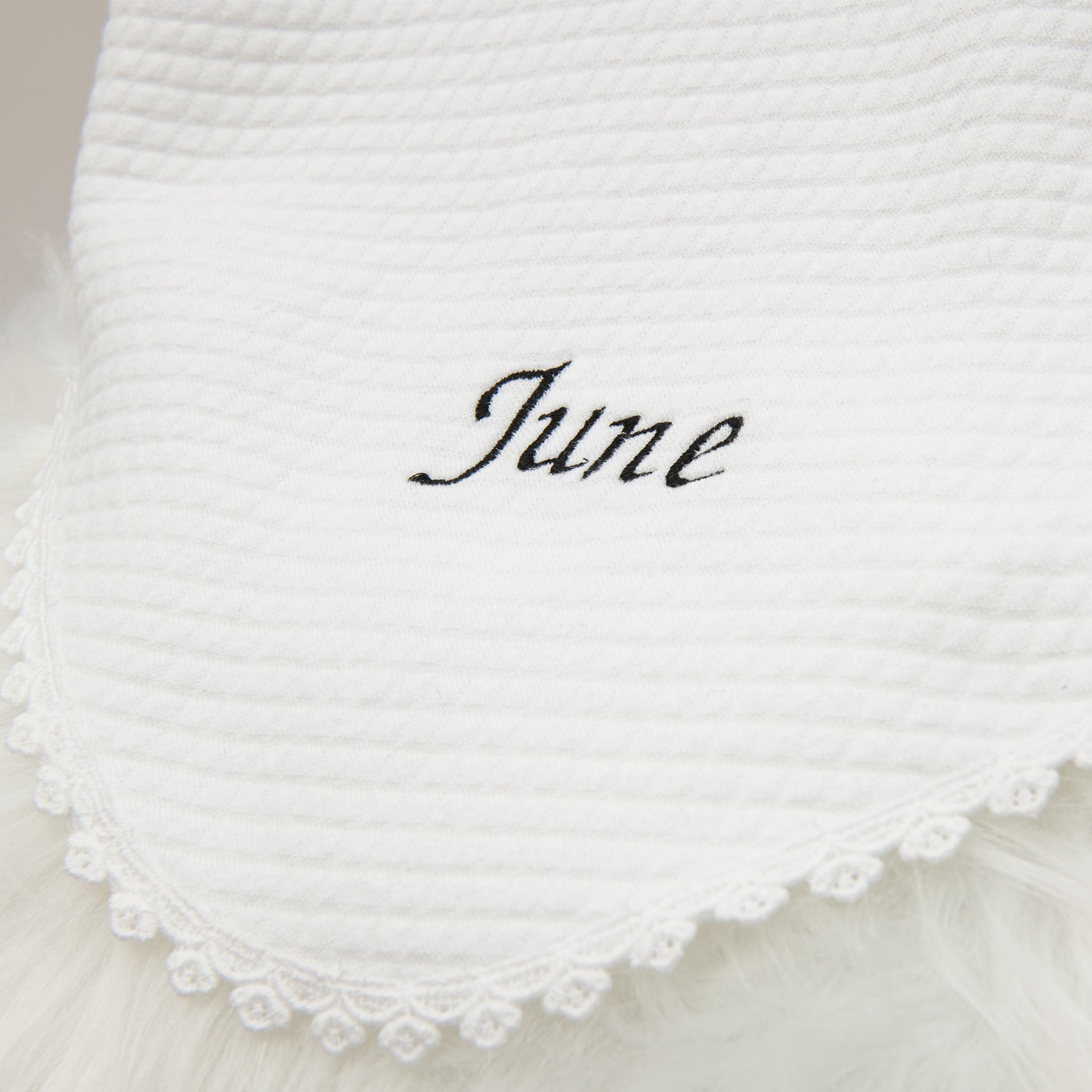 Close-up of a stylish white baby blanket with the name June embroidered in elegant black script, accented by a detailed lace trim along the edge.
