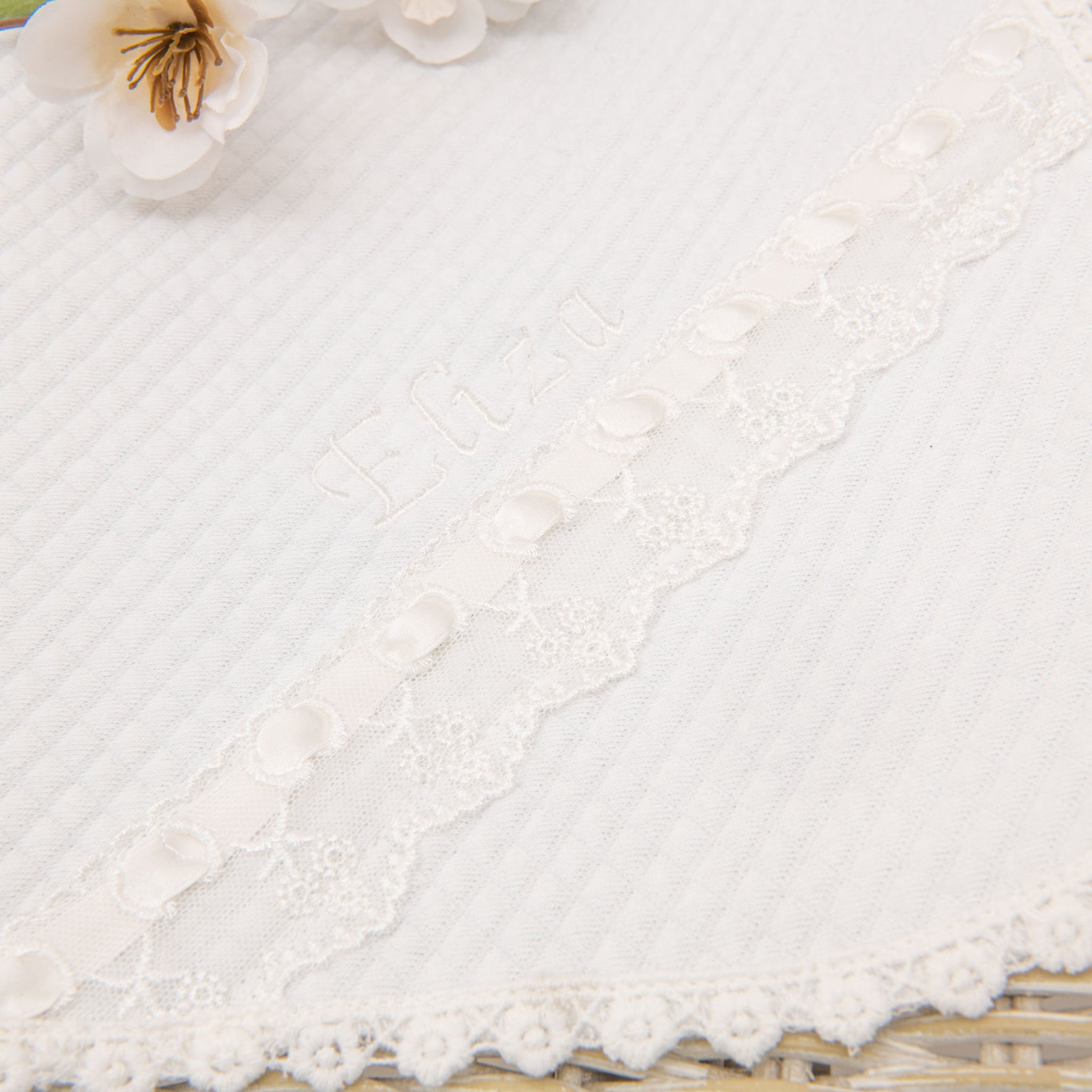 Close-up of a white textured fabric with embroidered flowers and floral lace trim. The product name "Eliza Personalized Blanket" is embroidered on the fabric, and there are decorative buttons along the lace edge. This handmade blanket appears to be a part of a baby blanket or similar delicate item.