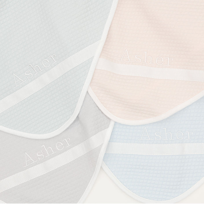 Flat lay photo of the four available colors of the Asher Personalized Blanket, including grey, pink, powder blue, and soft teal. The blanket is made out of a 100% Quilted Cotton and features an embroidered name on the corner of the blanket.