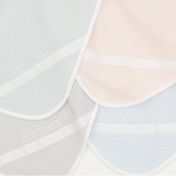 Flat lay photo of the four available colors of the Asher Personalized Blanket, including grey, pink, powder blue, and soft teal. The blanket is made out of a 100% Quilted Cotton and features an embroidered name on the corner of the blanket.