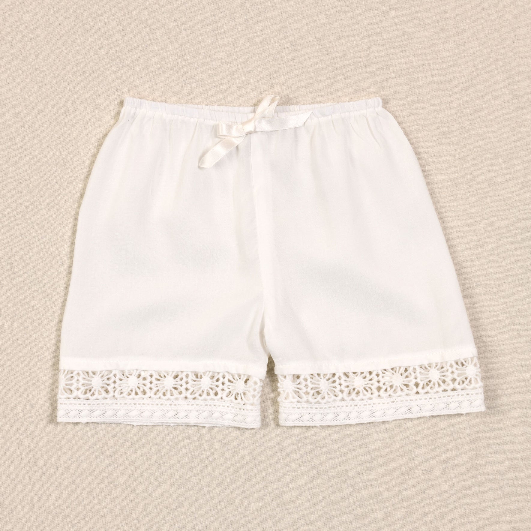 A pair of white, knee-length women's shorts displayed on a beige background. The shorts feature an elastic waistband with a decorative bow and intricate cotton lace trim at the hem, reminiscent of the Adeline Lace Christening Gown & Bonnet.