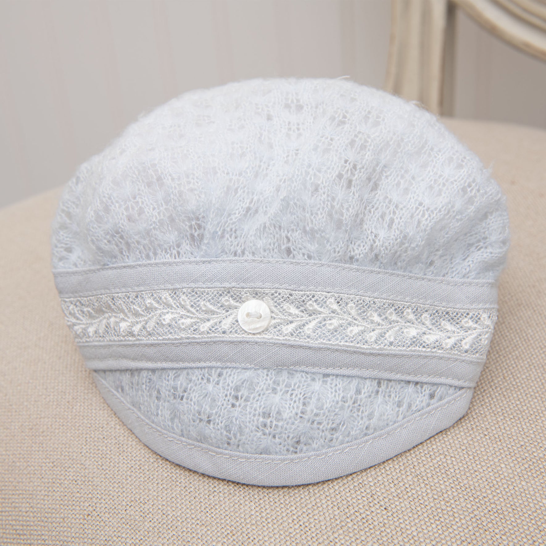 The Harrison Newborn Knit Hat, featuring a light gray color with a textured knit pattern, ivory Venice lace detailing, and a decorative button at the center front, is placed on a beige surface.