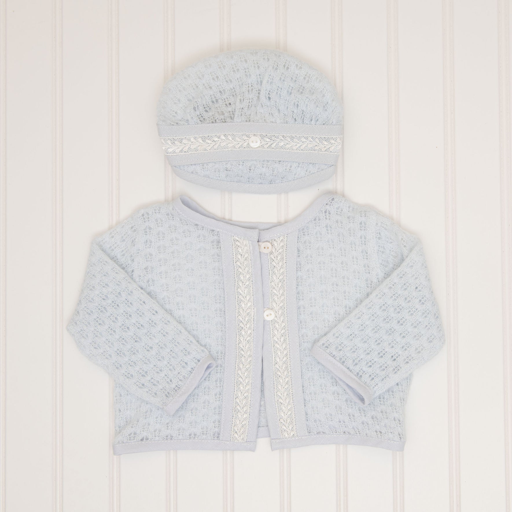 A light blue knit Harrison Romper Accessory Bundle is laid flat against a white background. The set includes a cardigan that features a textured pattern with two buttons and decorative trim along the edges, accompanied by a coordinating hat with matching trim, perfect for adding a delicate touch to any special occasion.