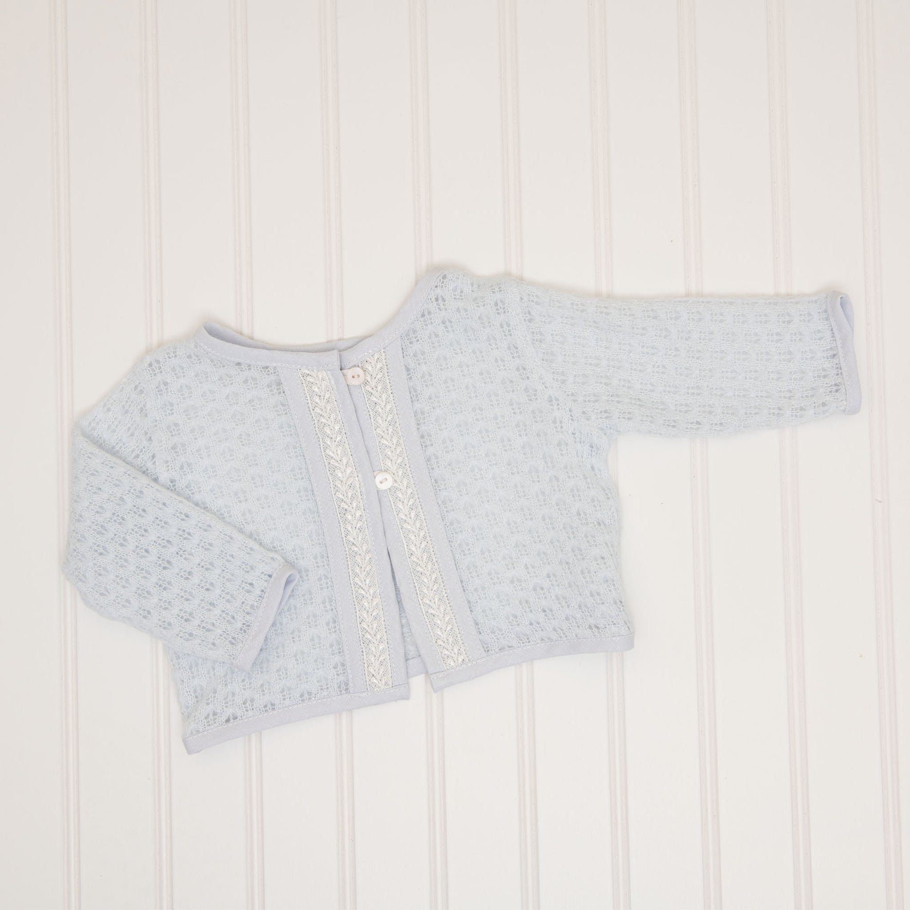 The Harrison Newborn Sweater, a soft blue knit sweater featuring a textured pattern and two buttons on the front, is laid flat on a white, subtly striped background.