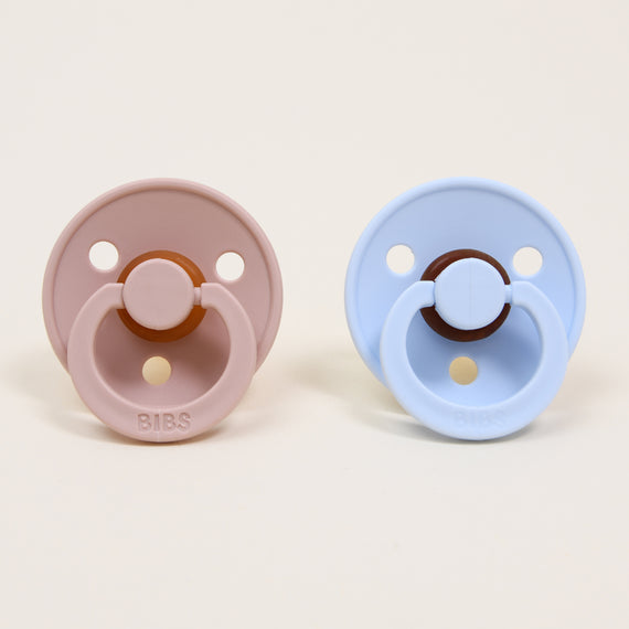 Displayed side by side are two round pacifiers from the Bibs Pacifier Set in Blush & Baby Blue, expertly crafted with natural rubber. The left one boasts a pink hue, while the right is adorned in light blue. Each pacifier features a central rubber nipple and a circular shield with two holes, and proudly bears the embossed word "BIBS" on its shield for self-soothing comfort, reflecting their Danish origins.