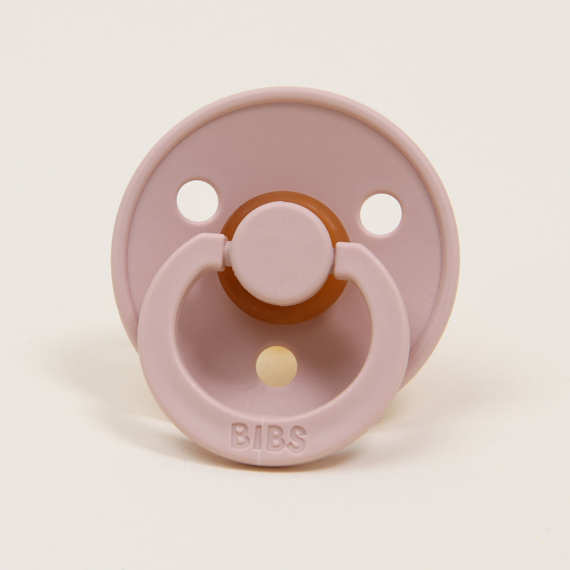 Displayed against a plain, light-colored background is a blush natural rubber baby pacifier featuring a circular shield with two holes and a ring handle. This pacifier, crafted in Denmark, has "BIBS" embossed on the handle and is part of the Bibs Pacifier Set | Dark Oak & Blush. It is perfect for helping little ones self-soothe.