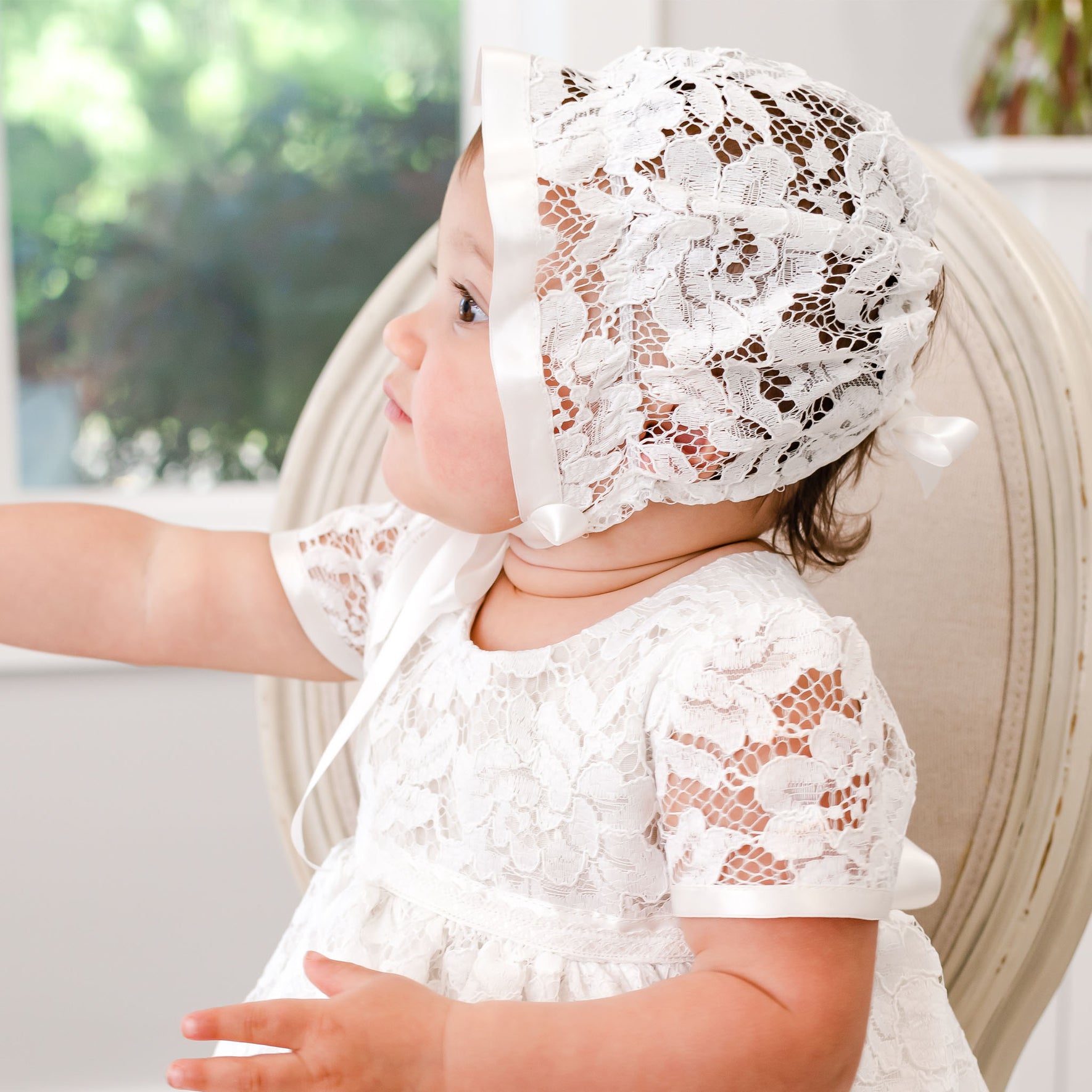 A baby with light skin wearing a Rose Christening Gown & Bonnet sits in a chair, gazing to the side in a brightly lit room.