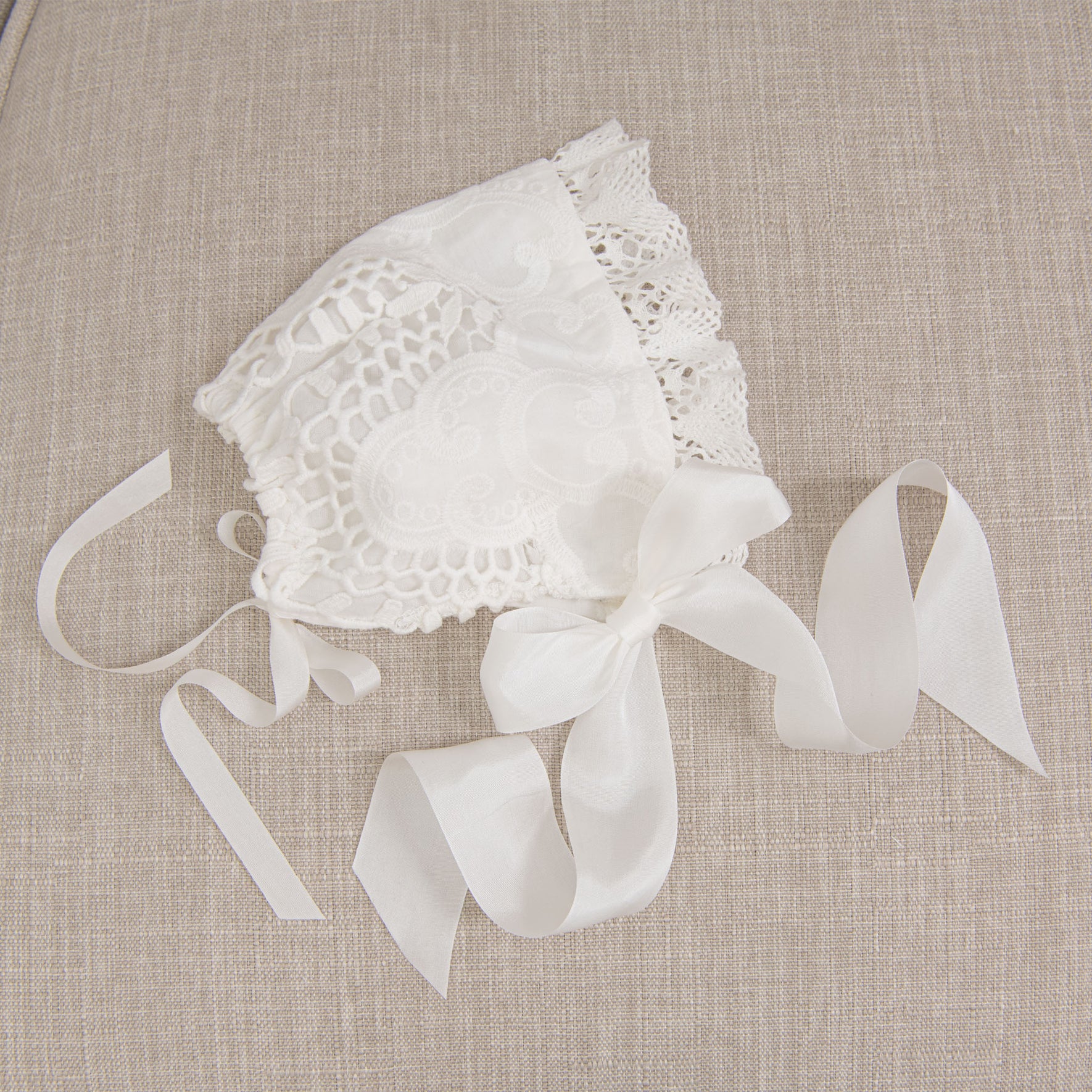Flat lay of the Lily Christening Bonnet resting on a beige fabric surface, emphasizing the delicate light ivory cotton lace, intricate edge lace, and soft ivory silk ribbons.