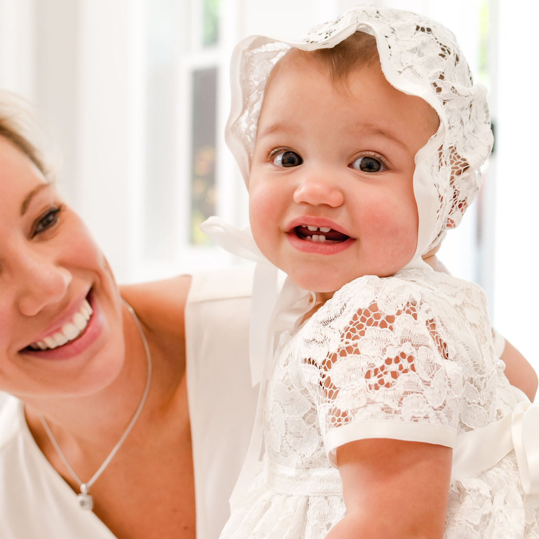 A joyful baby in a Rose Christening Gown & Bonnet smiles widely, held by a smiling woman with blonde hair. They are both indoors with a bright, airy background.