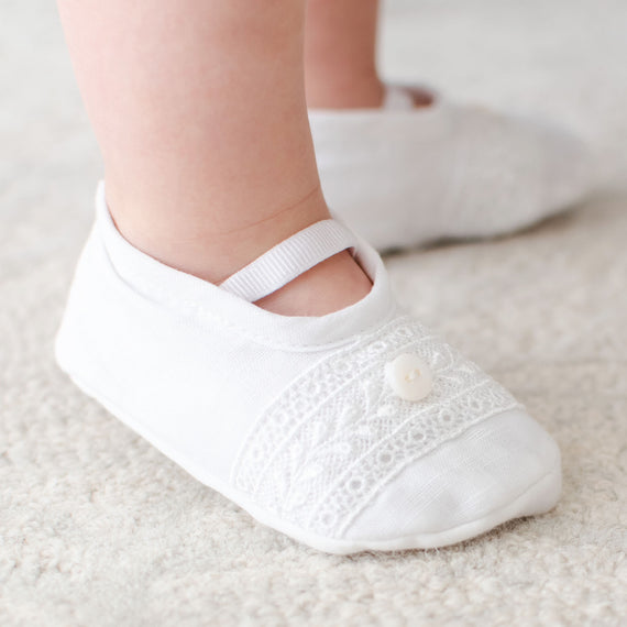 A close-up of a baby's feet wearing Oliver Booties, made from a linen/cotton blend and embellished with Venice lace and a decorative button. The shoes have a strap across the top and are placed on a textured, light-colored surface.