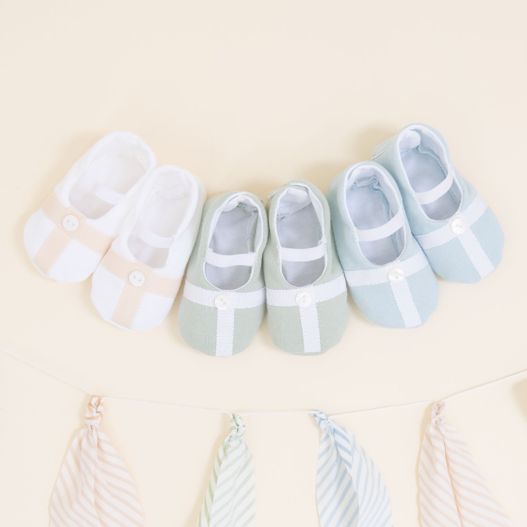 Flat lay photo showing the top detail of three pairs of Theodore Booties, including blue, green, and a white/tan color. The booties are made from a 100% French Terry Cotton with a Grosgrain Trim (with button detail) and soft elastic strap.