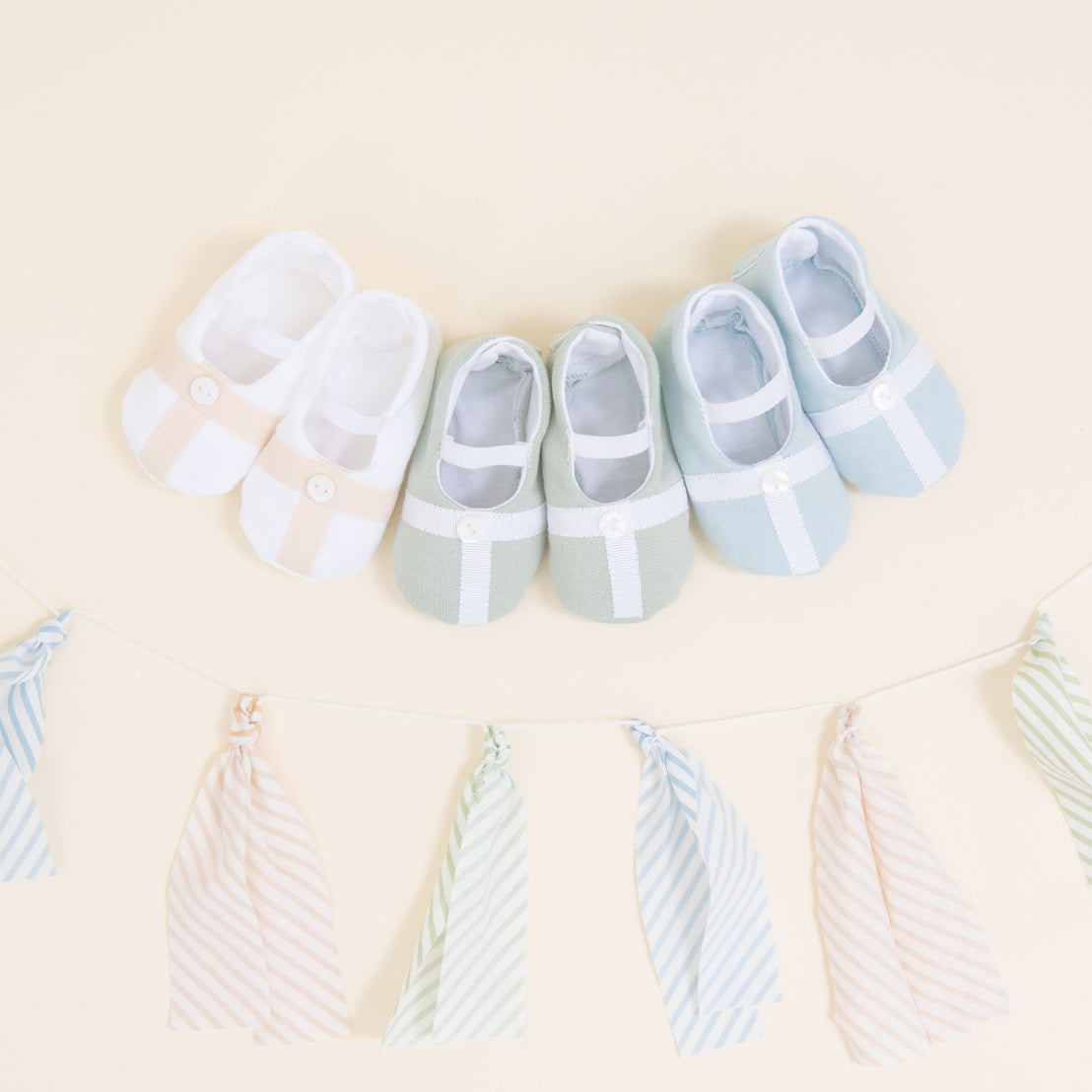 Flat lay photo showing the top detail of three pairs of Theodore Booties, including blue, green, and a white/tan color. The booties are made from a 100% French Terry Cotton with a Grosgrain Trim (with button detail) and soft elastic strap.