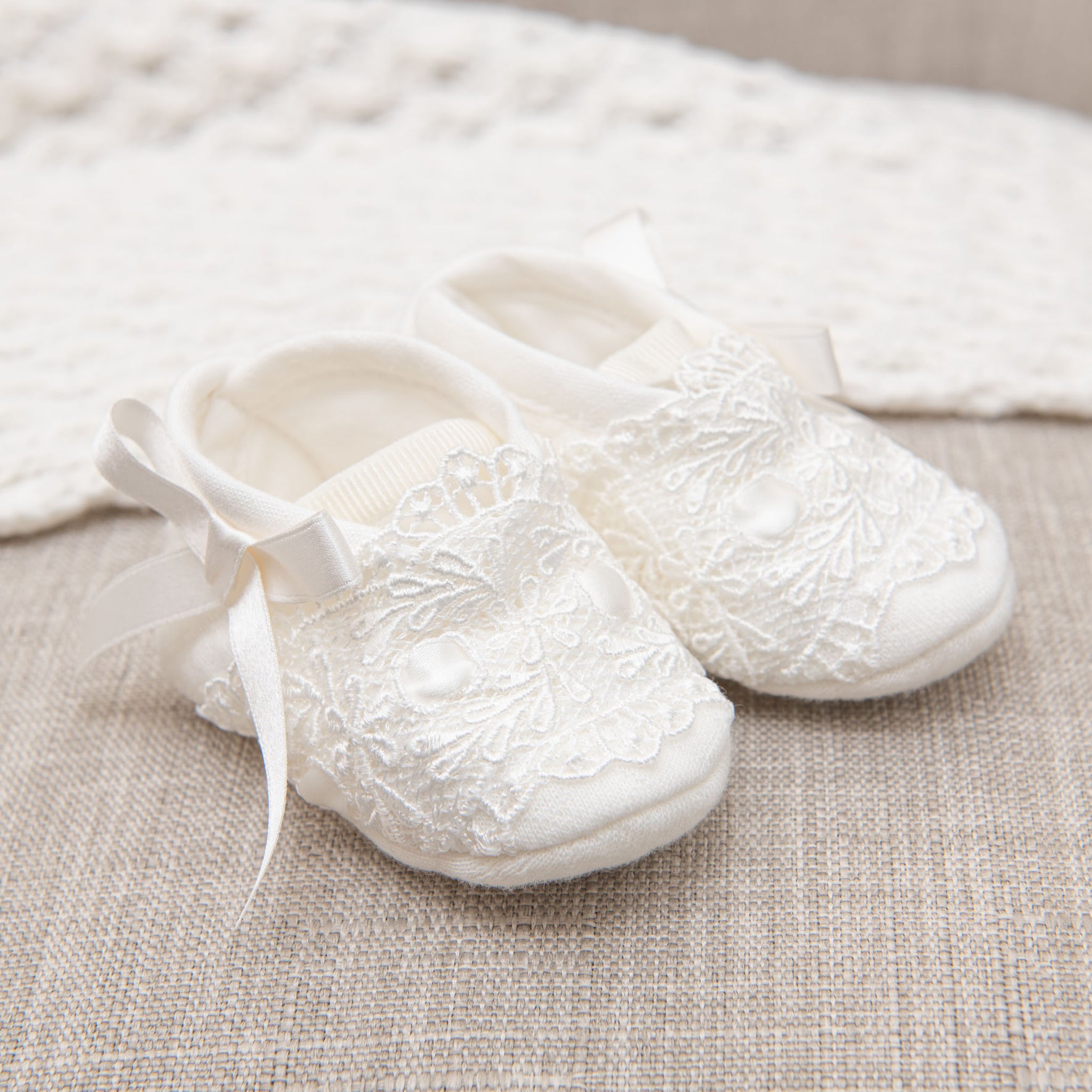 A pair of white baby shoes, adorned with delicate lace detailing and small bows, rests on a beige textured surface. Nearby, a knitted white blanket adds to the elegance, creating the Madeline Accessory Bundle - Save 15%, a charming newborn set perfect for welcoming little ones.