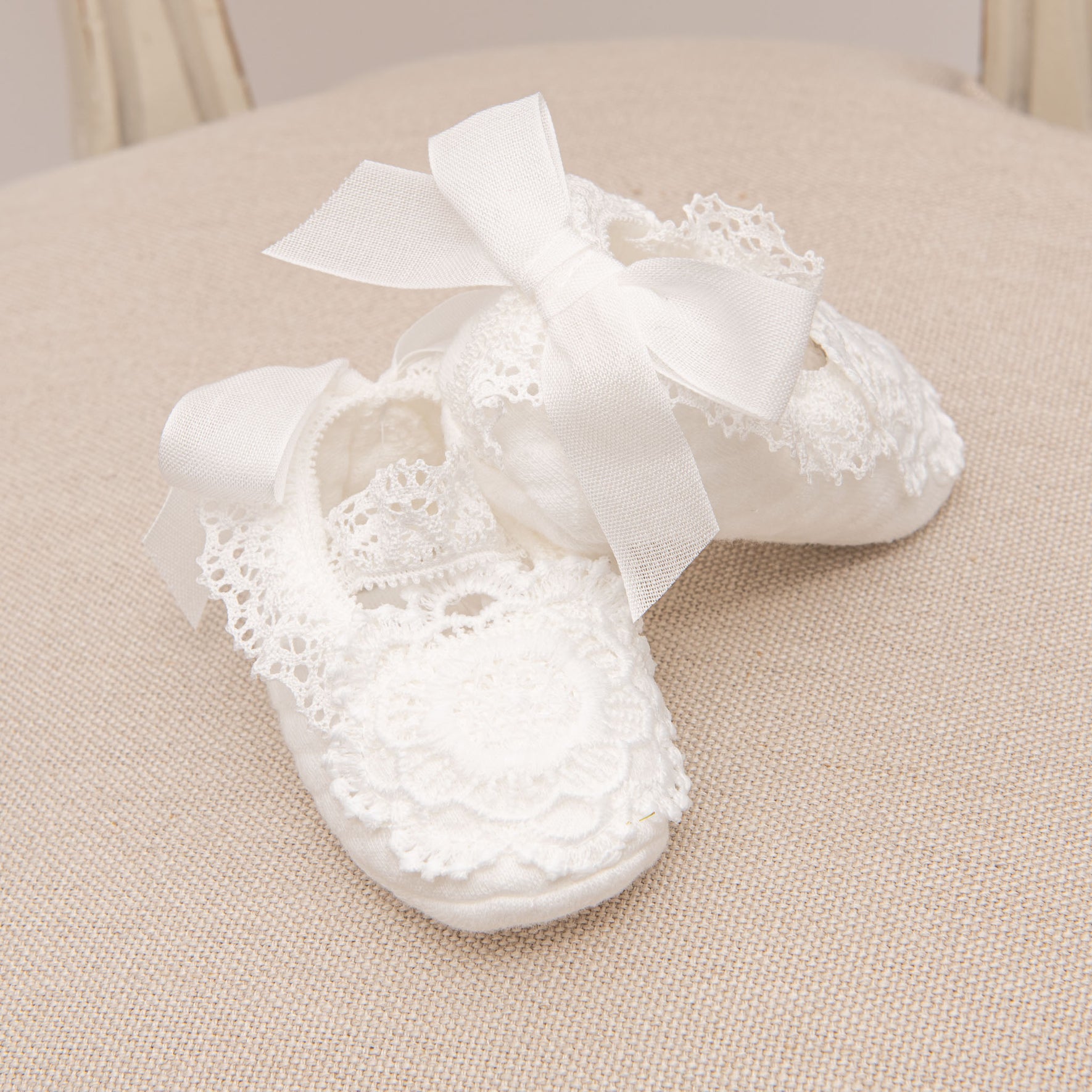 Poppy Christening Booties with intricate floral lace and satin ribbon ties, arranged neatly on a beige fabric surface.