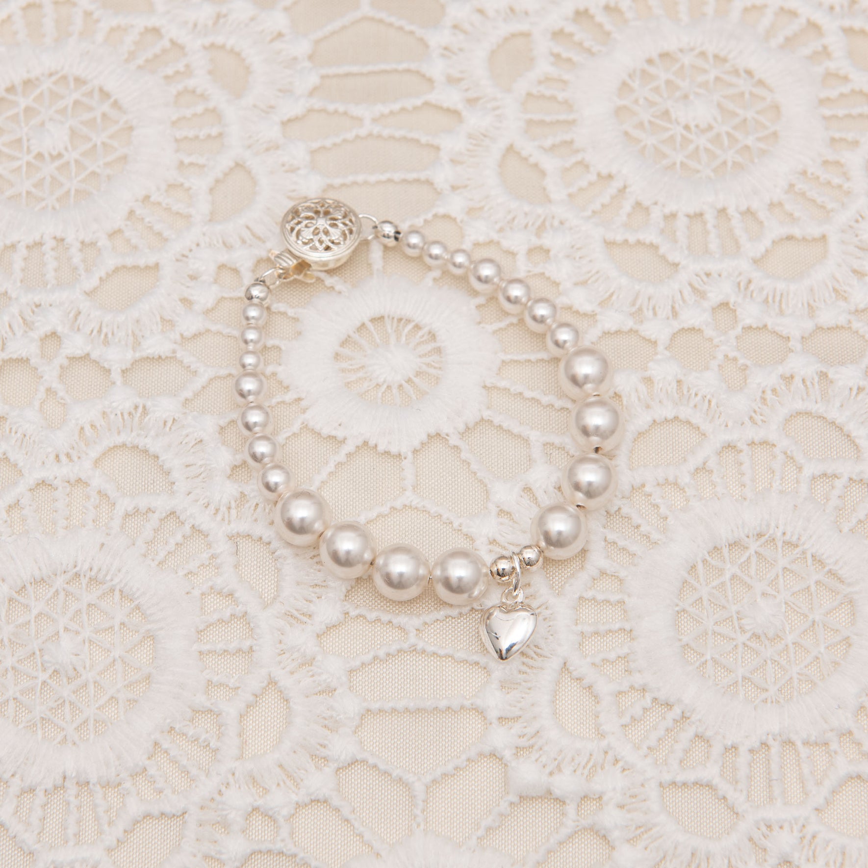 Close up of the White Luster Pearl Bracelet against a lace background. The bracelet features a silver heart charm, emphasizing its lustrous pearls and refined craftsmanship.