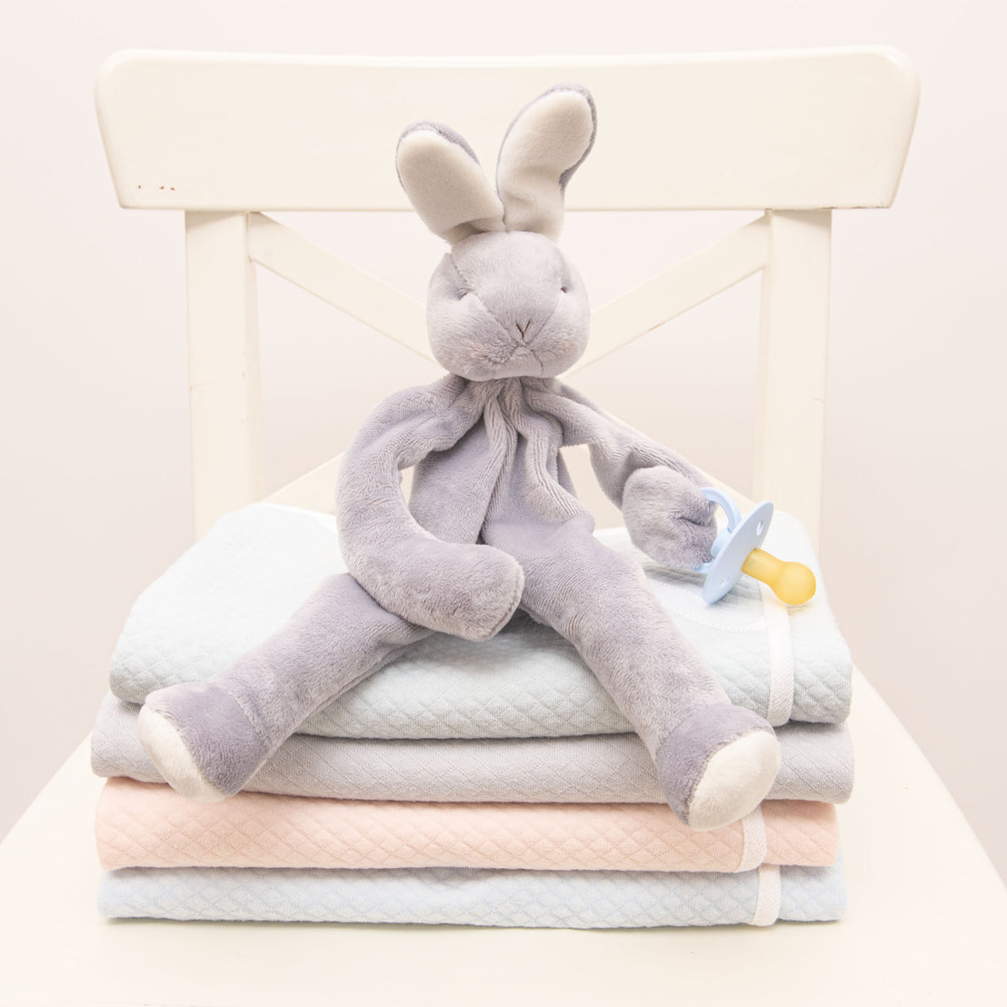 Flat lay photo of the Asher Silly Bunny Buddy, a soft stuffed animal bunny (made from a soft velour in grey) that holds on to a pacifier.
