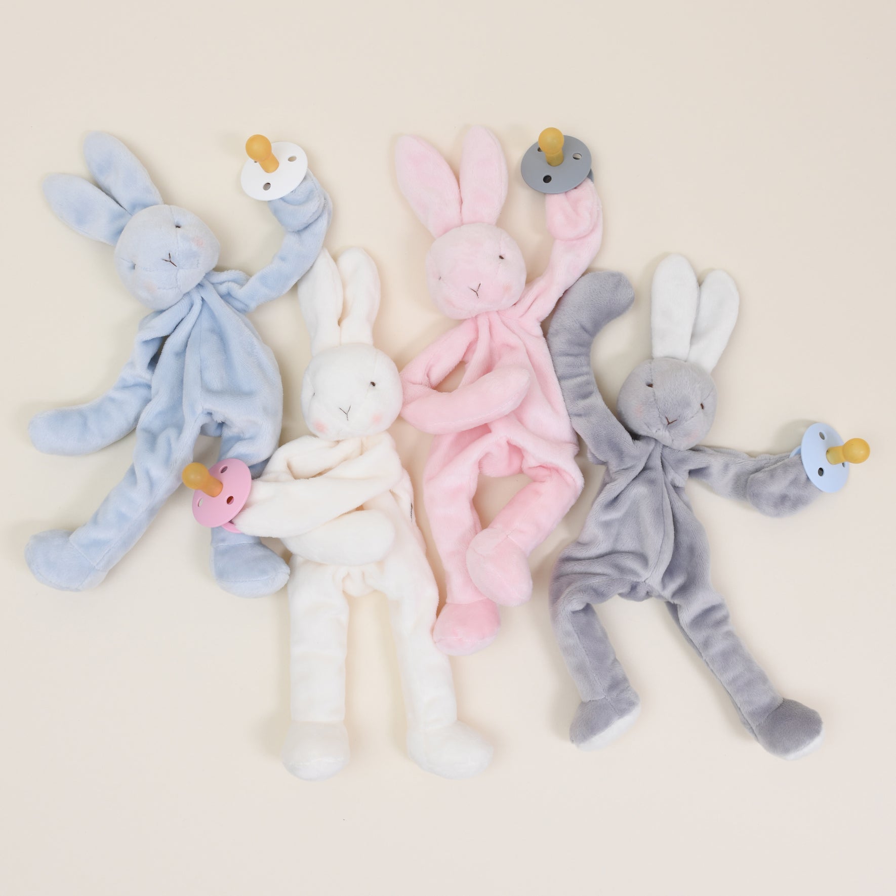 Four Silly Bunny Buddy Pacifier Holders in blue, white, pink, and gray are arranged on a neutral background. Each baby-safe rabbit plush features floppy ears and limbs while holding a matching colored pacifier with a clip: blue, white, pink, and gray.