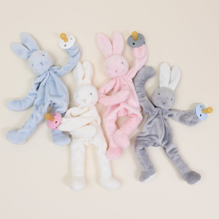 Four Silly Bunny Buddy Pacifier Holders in blue, white, pink, and gray are arranged on a neutral background. Each baby-safe rabbit plush features floppy ears and limbs while holding a matching colored pacifier with a clip: blue, white, pink, and gray.