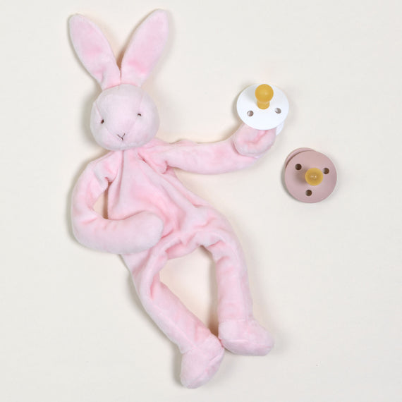 A Pink Silly Bunny Buddy gently cradles a pacifier with a soft pink exterior and yellow nipple in one hand. Close by, another pacifier with a brown finish and beige nipple rests against the light background. Designed with baby safety in mind, this charming toy ensures your little one's essentials are kept close and secure.