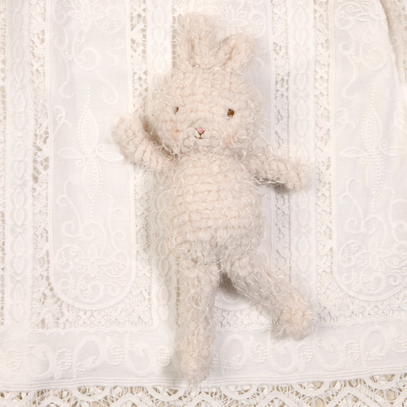 The Adeline Bunny Friend, a petite crocheted rabbit toy featuring soft cream fur and a fluffy texture, rests on a white lace-patterned fabric background. With pink cheeks and a simple stitched face, this plush bunny makes for an adorable baby shower gift.