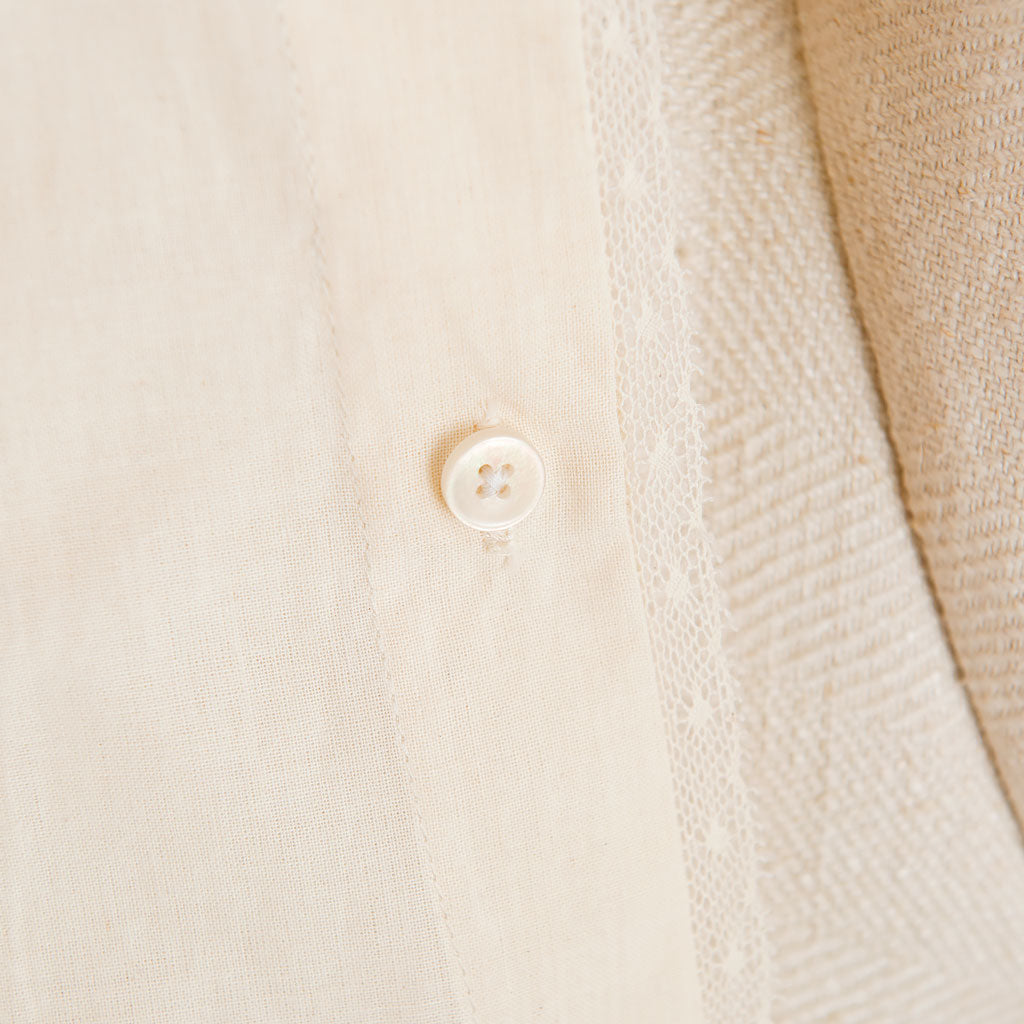 A close-up of a white button on the cream-colored Muslin Keepsake Garment Bag, made from 100% cotton muslin with a subtle pattern. The fabric surrounding the button features stitching details that enhance texture, resembling delicate lace edging. The focus is on the button and the adjacent cloth.