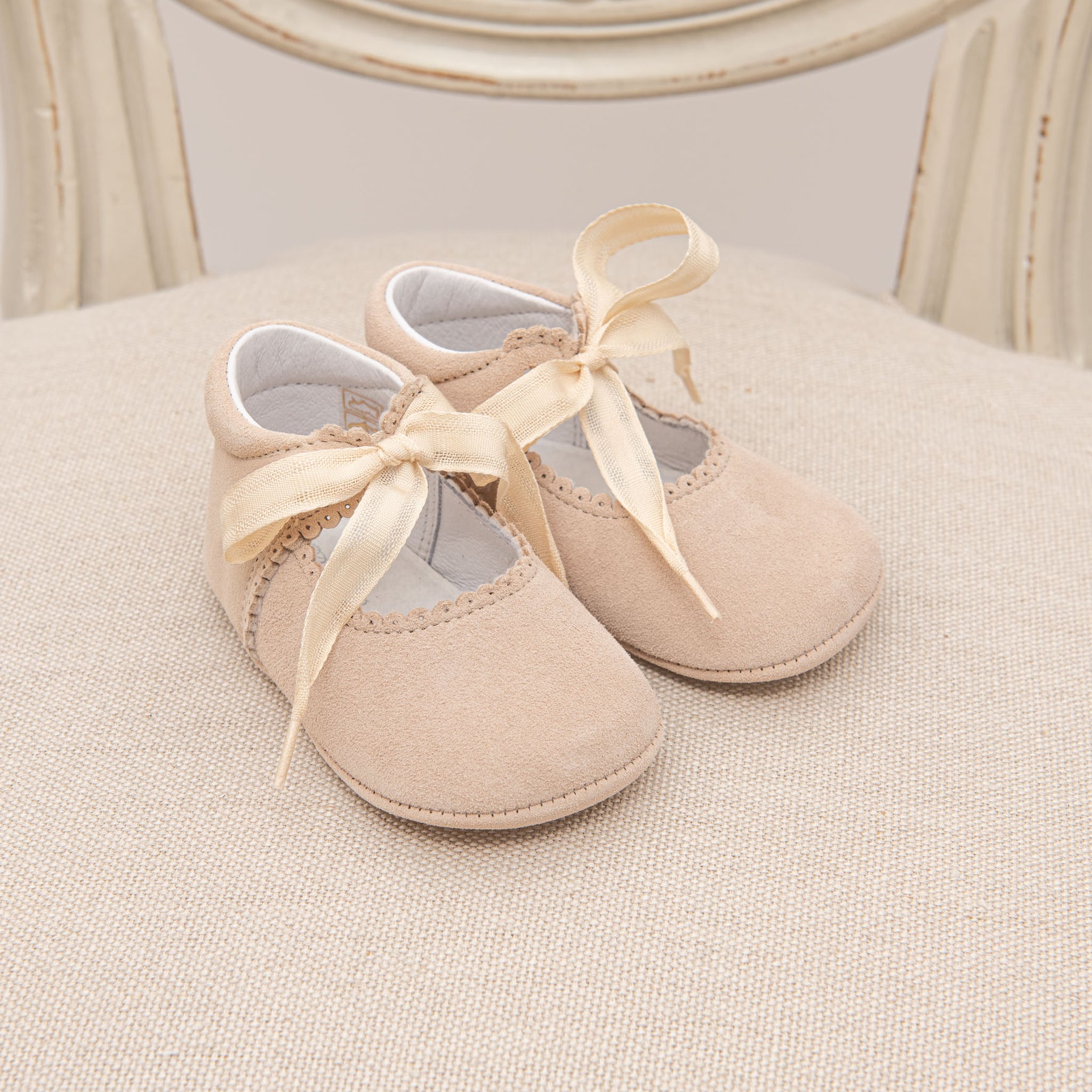 A pair of small, beige Kristina Tie Mary Janes with satin ribbon bows are placed on a cream-colored fabric surface. The handmade baby shoes feature scalloped edges and a soft, cushioned feel. A wooden chair with a beige finish is partially visible in the background.
