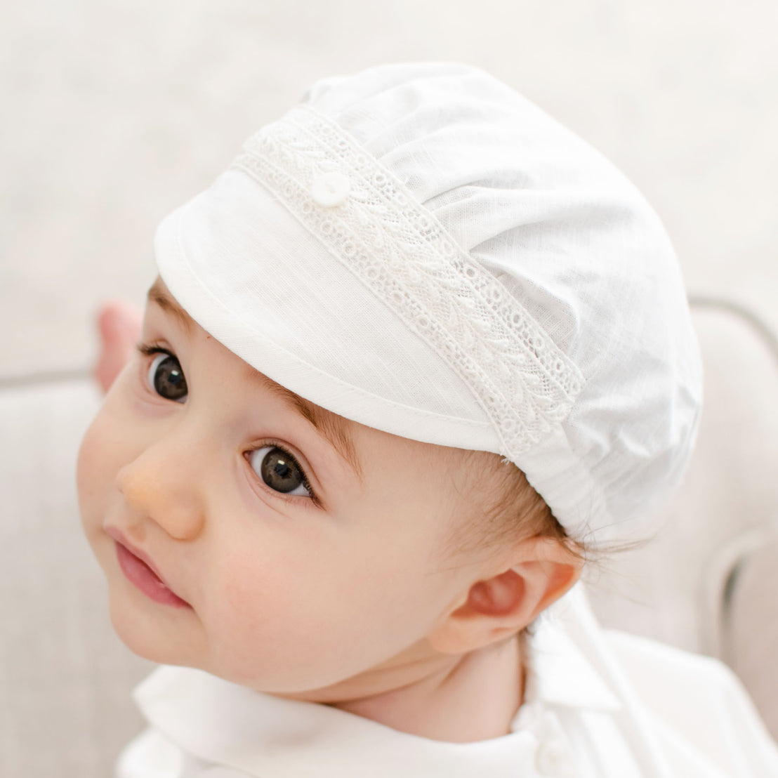 Baby boy wearing a white Oliver Linen Cap. The hat is made from linen and features a Venice lace across the front and a button for added detail.