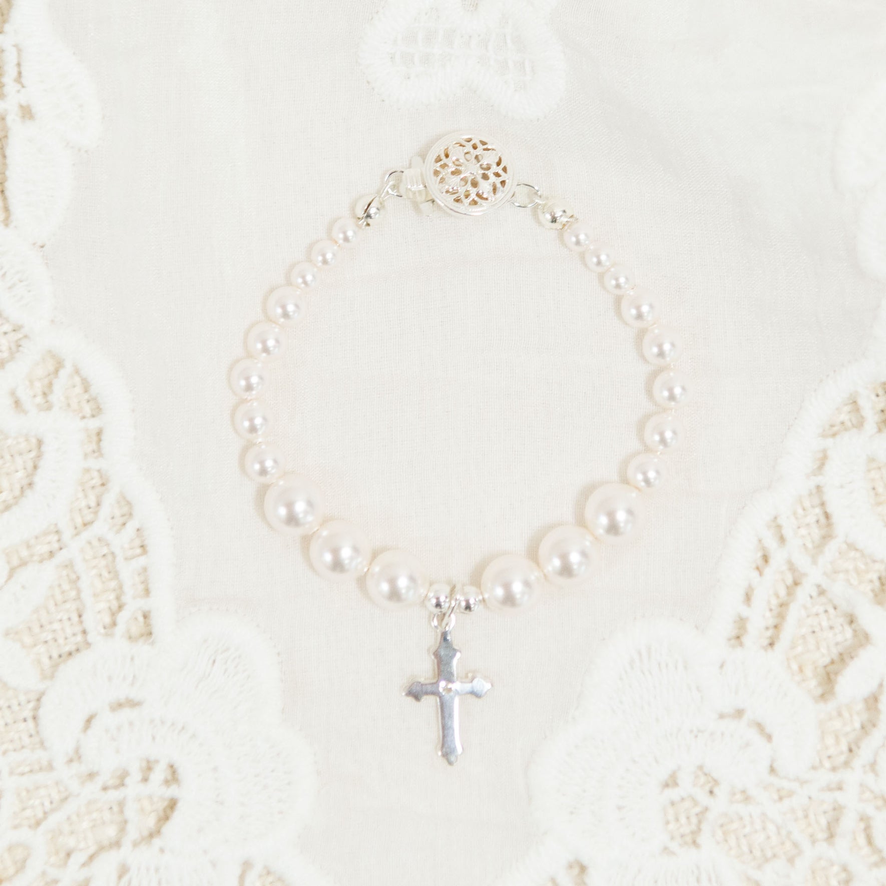 The White Luster Pearl Bracelet with Silver Cross is elegantly displayed on a white lace fabric background, making it an ideal Christening gift. The bracelet boasts a decorative circular clasp, enhancing its intricate beauty.
