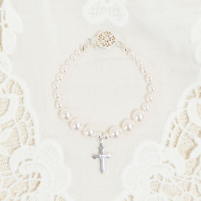 The White Luster Pearl Bracelet with Silver Cross is elegantly displayed on a white lace fabric background, making it an ideal Christening gift. The bracelet boasts a decorative circular clasp, enhancing its intricate beauty.