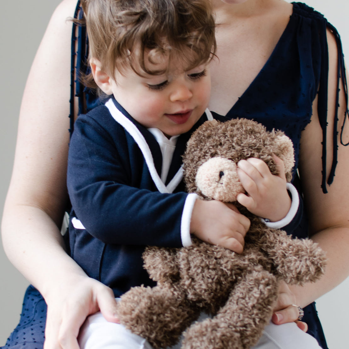 Smiling baby boy sitting on his mother's lap. He is wearing the Elliott 3-Piece Suit, including the jacket, pants, and onesie. He is also holding the Cubby Bear.