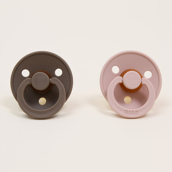 The Bibs Pacifier Set from Denmark features two pacifiers displayed side by side. The one on the left, crafted from natural rubber and called Dark Oak, is a dark brown shade, while the right one is Light Pink, known as Blush. Both have a simple round design with a looped handle, ideal for helping babies self-soothe against a plain background.