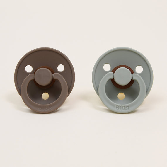 Two Bibs pacifiers from the Dark Oak & Sage set are displayed against a plain background. The classic design features the pacifier on the left in a dark oak shade and the one on the right in sage. Both have round shields with two ventilation holes and a ring handle, ideal for self-soothing.