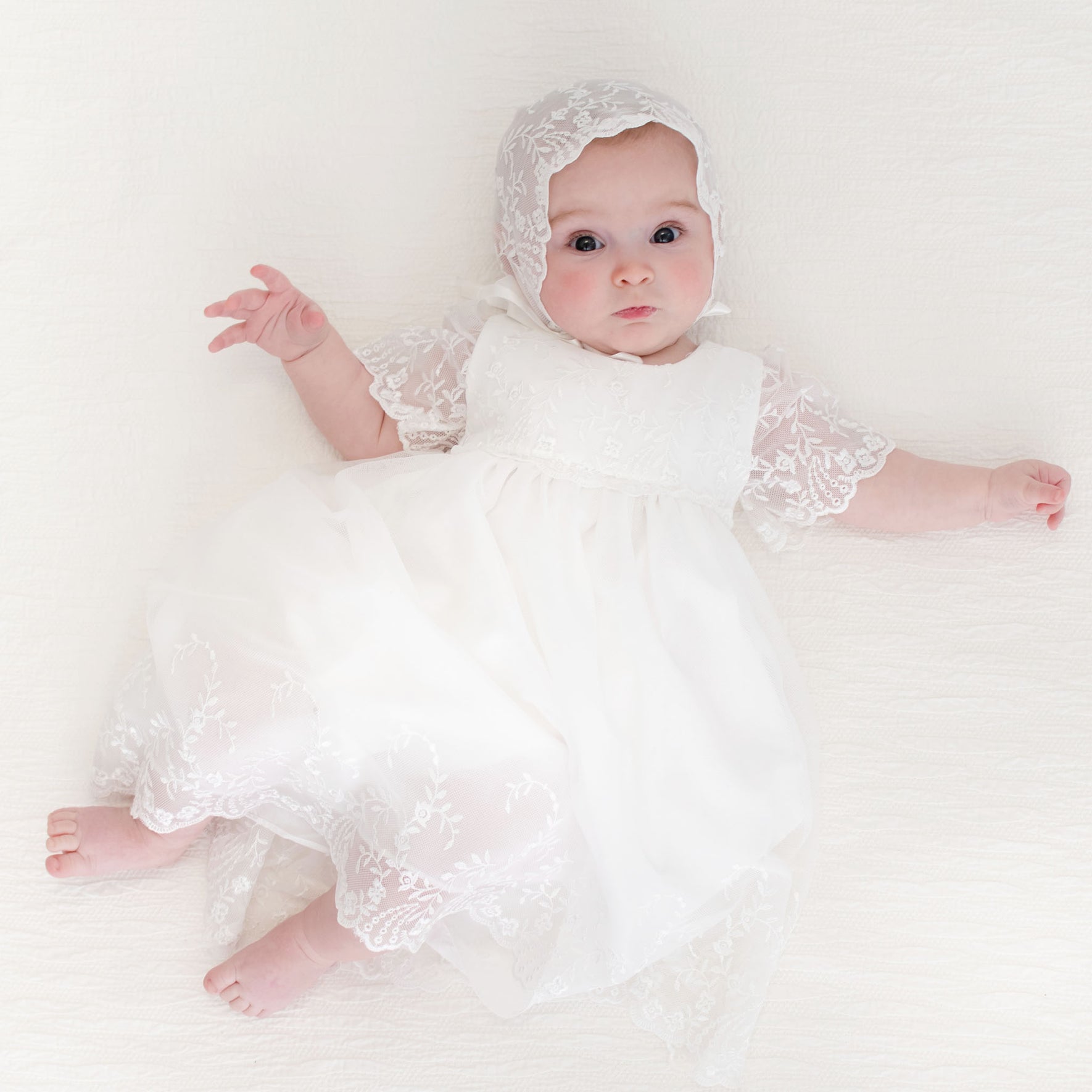Baby girl dressed in the Ella baptsim dress and Ella Lace Bonnet, gazing curiously at the camera with open arms.