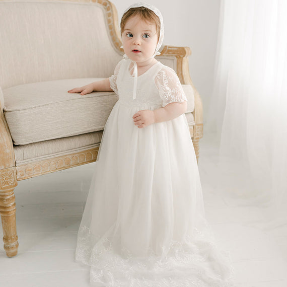 baby girl standing by chair wearing Ella christening gown 