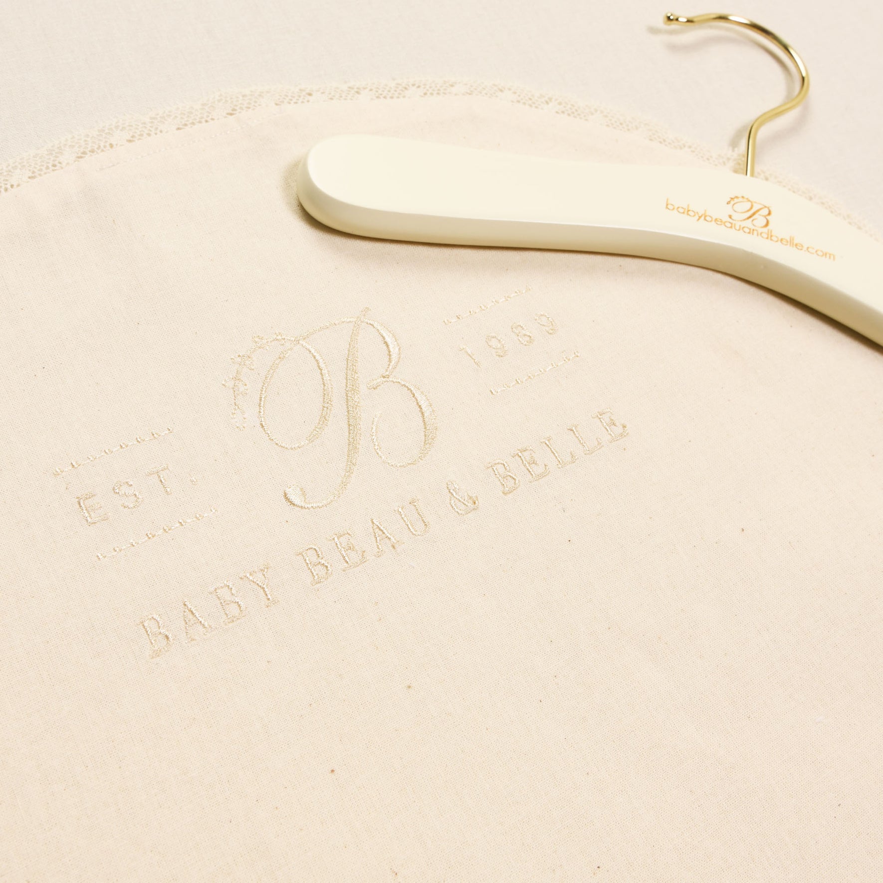 This Muslin Keepsake Garment Bag, crafted from 100% cotton muslin, showcases a subtle embroidery of "Baby Beau & Belle" and "EST 1989" in cream-colored fabric. The elegant design includes a nearby hanger featuring the brand's website, making it perfect for stylish storage.