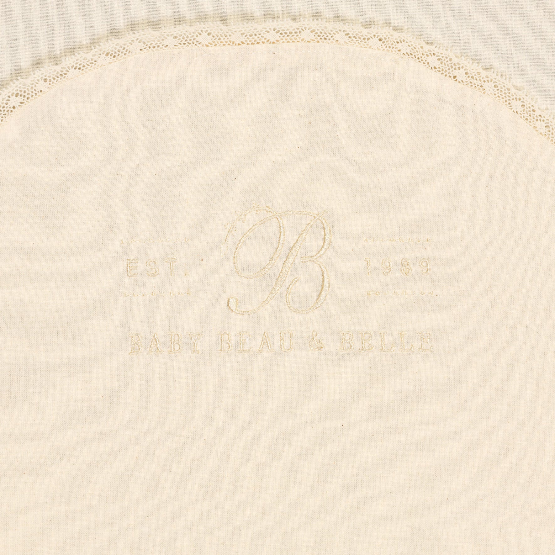 A cream-colored Muslin Keepsake Garment Bag, featuring a delicate lace edging and crafted from 100% cotton muslin. The text "Baby Beau & Belle" is embroidered in the center, along with "Est. 1989" and the letter "B".
