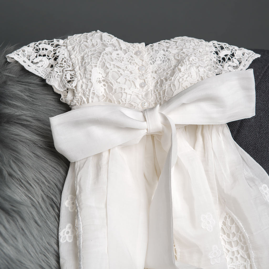 The Grace Christening Gown & Bonnet is showcased on a gray fabric surface, featuring intricate lace detailing on the bodice, with a large white bow adorning the back. The dress has a soft, flowing skirt and is paired with a matching lace Christening Bonnet. The background includes a section of light gray fur fabric.
