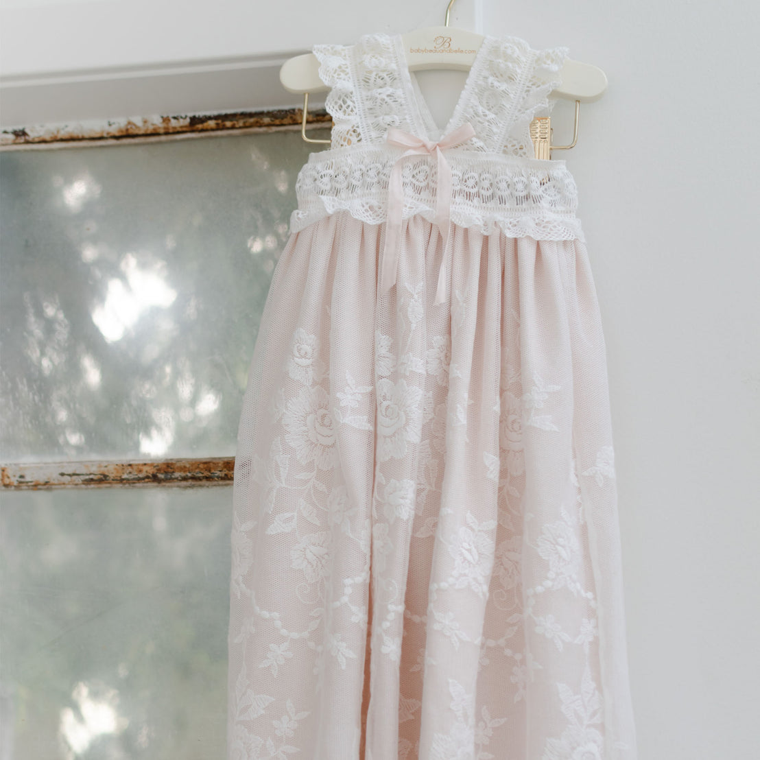 A delicate pink and white Charlotte Christening Gown & Bonnet hangs on a wooden hanger against a white wall, reflecting soft light. The dress features intricate lace patterns and a flowing skirt.
