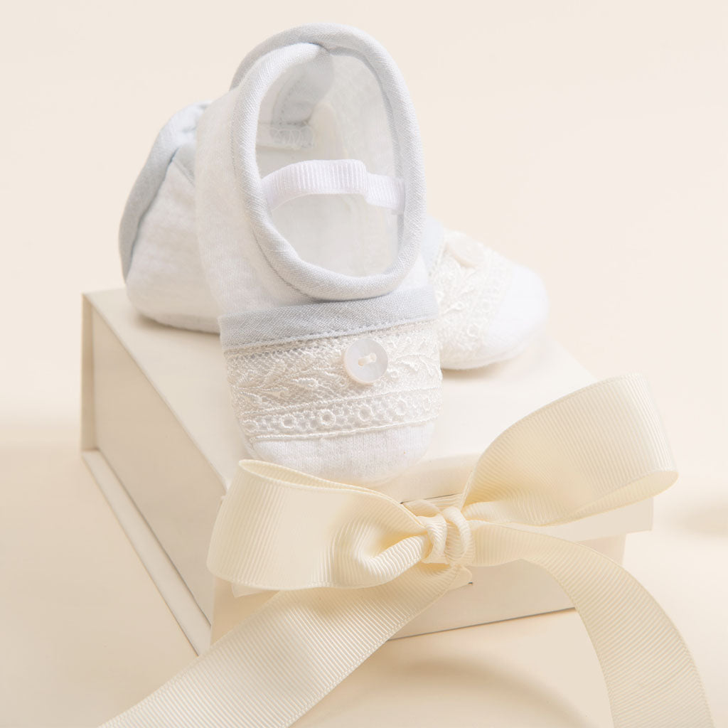 A pair of white baby booties with lace details is elegantly displayed on Baby Beau & Belle's "Gift Wrap | Mini Gift Box with Bow." The box features a keepsake-quality cream ribbon tied into an elegant bow, set against a soft beige background.