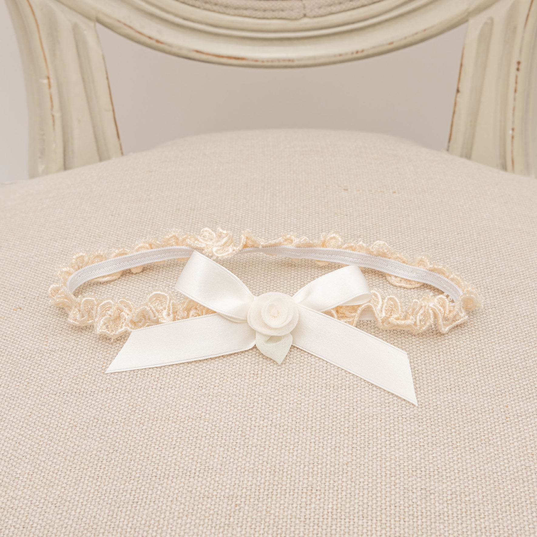 The Kristina Accessory Bundle, which saves you 15%, includes an elegant beige lace garter adorned with a white satin bow and rosette, beautifully enhancing the delicate charm of a baby girl Christening outfit as it graces a beige cushioned chair.