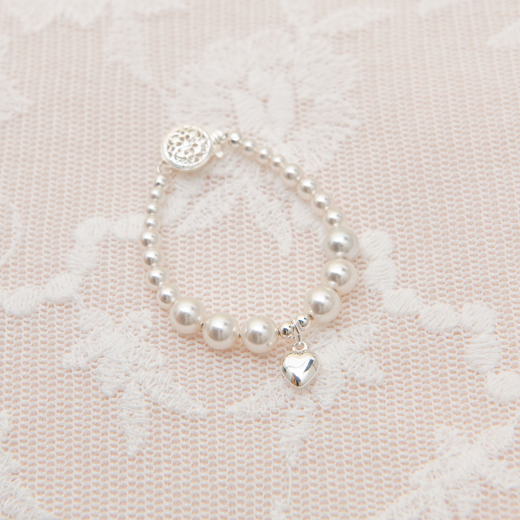 The White Luster Pearl Bracelet with Silver Heart Charm, featuring smooth round beads and a small sterling silver heart-shaped accent, is elegantly displayed on a delicate lace fabric background.