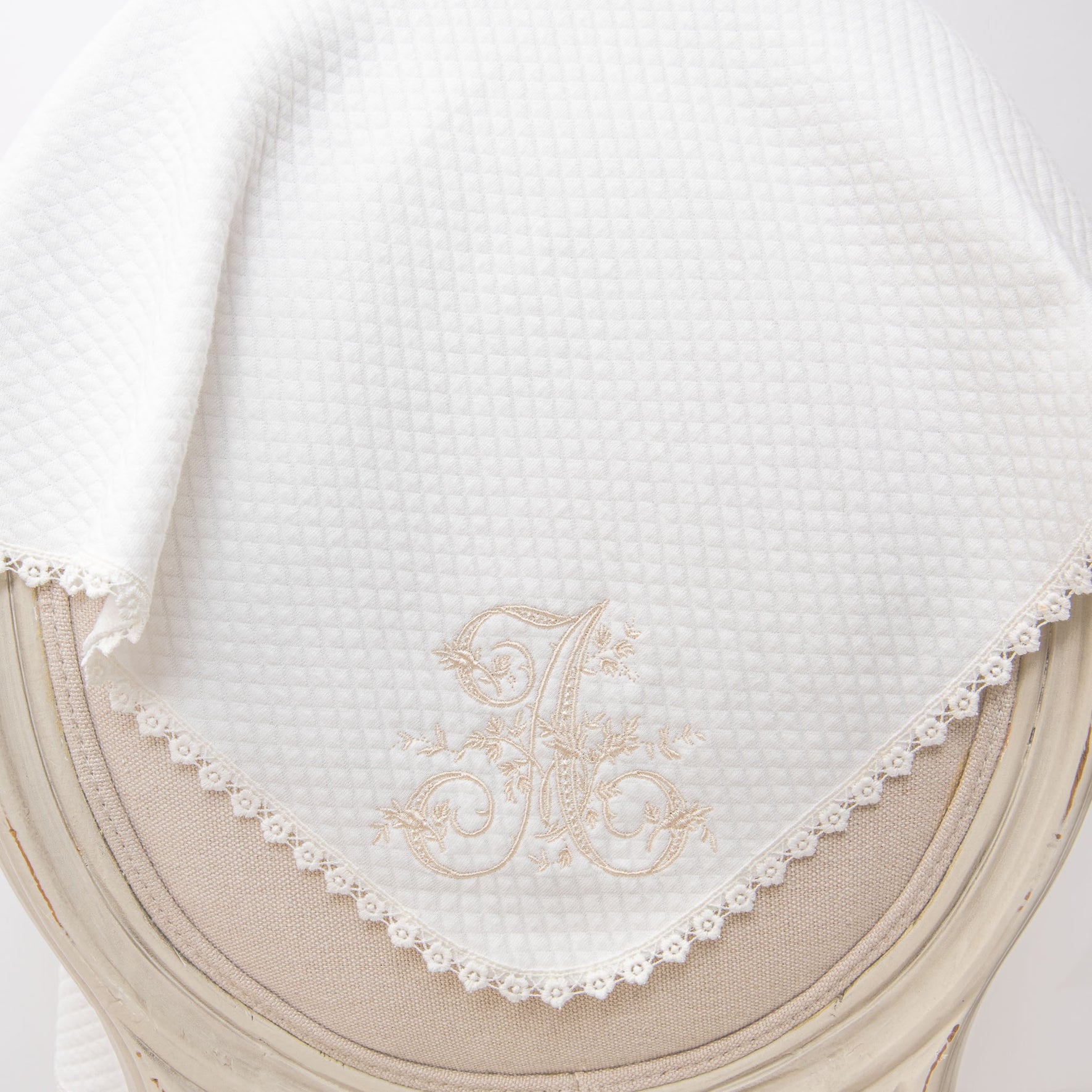The Initial Blanket, a white quilted cotton baby blanket featuring a lace-trimmed edge, drapes elegantly over a beige upholstered chair. Adorned with an ornate embroidered letter "A" and light beige floral accents, it offers a charming touch of personalized embroidery.
